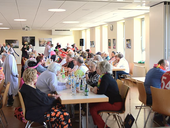 In einem Saal sitzen an langen Tischen zahlreiche bunt maskierte Seniorinnen und Senioren und im Hintergrund sind man einige Tanzpaare. 