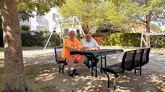 Zwei Männer sitzen an einer neuen Sitzgruppe auf dem Spielplatz „Am Schafgraben“, im Hintergrund sind die erneuerte Doppelschaukel sowie weitere Spielgeräte unter schattenspendenden Bäumen zu sehen.