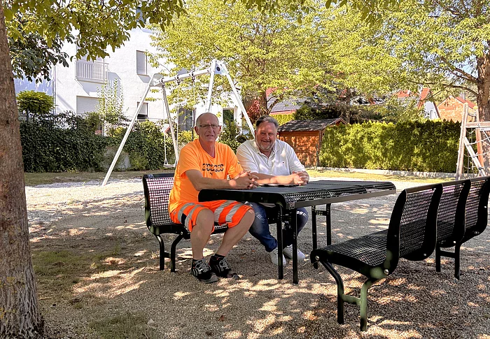 Zwei Männer sitzen an einer neuen Sitzgruppe auf dem Spielplatz „Am Schafgraben“, im Hintergrund sind die erneuerte Doppelschaukel sowie weitere Spielgeräte unter schattenspendenden Bäumen zu sehen.