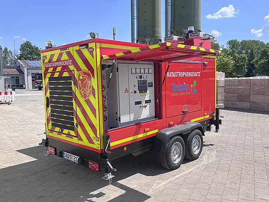 Ein mobiles rotes Notstromaggregat auf einem Anhänger steht auf dem Gelände eines Wasserwerks. Die Seitenklappe ist geöffnet und gibt den Blick auf die technische Steuerungs- und Schaltanlage frei. Auf dem Fahrzeug sind reflektierende Warnmarkierungen sowie die Aufschrift „Katastrophenschutz Teublitz“ zu sehen.
