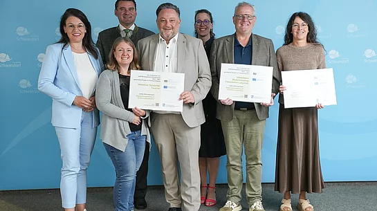 Gruppenfoto von sieben Personen bei der Übergabe mehrerer LEADER-Förderbescheide im Landratsamt Schwandorf. Im Vordergrund halten drei Personen große Fördertafeln in den Händen. Eine davon weist die Förderung für den „Inklusiven Spielplatz Teublitz“ aus. Die Gruppe steht vor einer blauen Wand mit dem Logo des Landkreises Schwandorf.