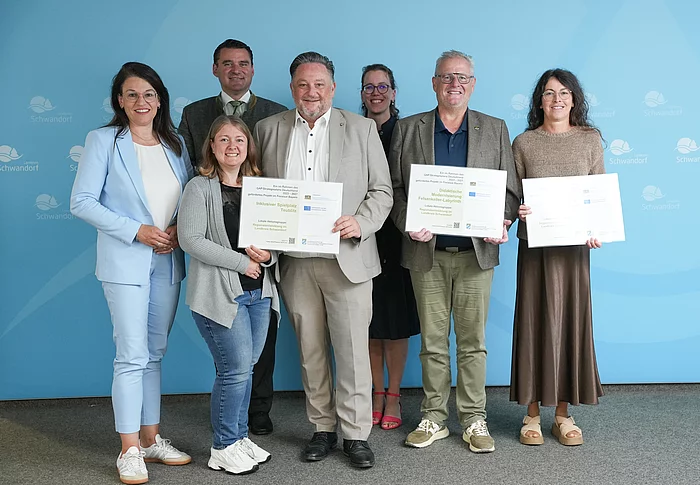 Gruppenfoto von sieben Personen bei der Übergabe mehrerer LEADER-Förderbescheide im Landratsamt Schwandorf. Im Vordergrund halten drei Personen große Fördertafeln in den Händen. Eine davon weist die Förderung für den „Inklusiven Spielplatz Teublitz“ aus. Die Gruppe steht vor einer blauen Wand mit dem Logo des Landkreises Schwandorf.
