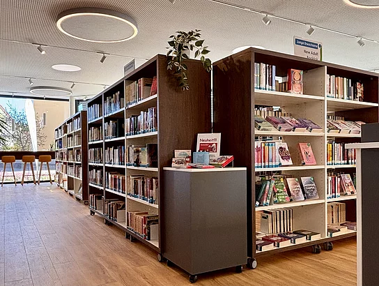 Mehrere Bücherregale mit Romanen und Neuerscheinungen stehen in einem warm beleuchteten Bereich der Bücherei.