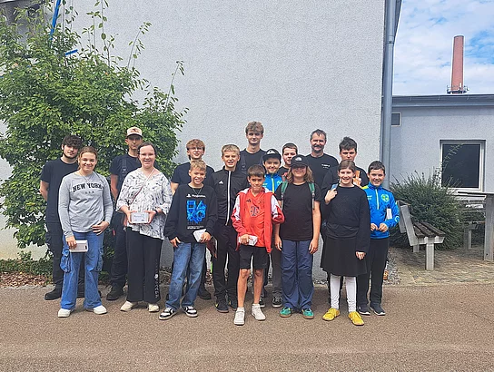 Eine größere Gruppe von Kindern, Jugendlichen und erwachsenen Betreuern steht für ein Gruppenfoto im Freien vor einem Betriebsgebäude. Einige Kinder halten kleine Schachteln oder ihre Arbeitsergebnisse in den Händen.