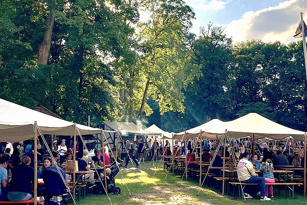 Mehrere große weiße Zelte stehen auf einer Wiese im Stadtpark. Davor sitzen zahlreiche Besucherinnen und Besucher an langen Bierbänken und genießen Speisen und Getränke. Zwischen den Zelten herrscht reges Treiben, während Sonnenstrahlen durch die Baumkronen fallen.