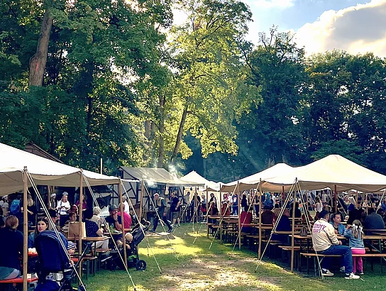 Mehrere große weiße Zelte stehen auf einer Wiese im Stadtpark. Davor sitzen zahlreiche Besucherinnen und Besucher an langen Bierbänken und genießen Speisen und Getränke. Zwischen den Zelten herrscht reges Treiben, während Sonnenstrahlen durch die Baumkronen fallen.