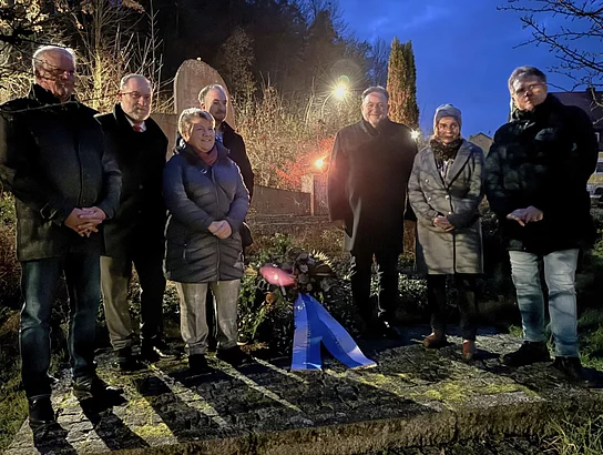 Eine kleine Gruppe von Menschen steht in der Dämmerung vor einem Gedenkstein und einem niedergelegten Kranz, beleuchtet von warmem Licht.