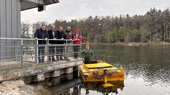 Mehrere Männer stehen auf einer Plattform an einem Gebäude am Seeufer, während daneben ein gelbes Mähboot im Wasser liegt und ein weiterer Mann darauf steht.