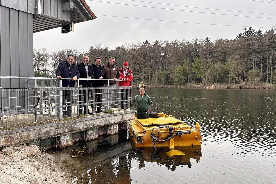 Mehrere Männer stehen auf einer Plattform an einem Gebäude am Seeufer, während daneben ein gelbes Mähboot im Wasser liegt und ein weiterer Mann darauf steht.