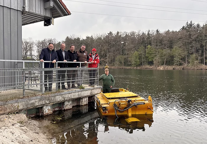 Mehrere Männer stehen auf einer Plattform an einem Gebäude am Seeufer, während daneben ein gelbes Mähboot im Wasser liegt und ein weiterer Mann darauf steht.