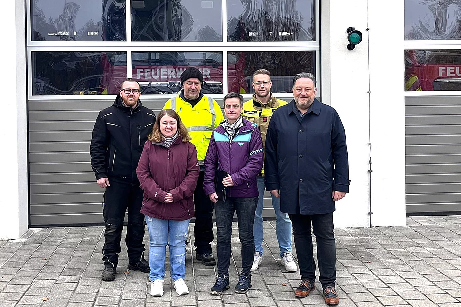 Auf dem Foto stehen sechs Personen – drei Männer und drei Frauen – lächelnd vor einem Feuerwehrgebäude mit grauen Sektionaltoren und der Aufschrift „FEUERWEHR“ auf den Fahrzeugen im Hintergrund.