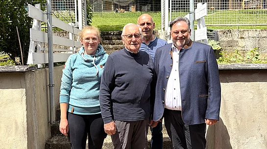 Vier Personen stehen bei sonnigem Wetter vor einer Treppe mit weißem Geländer und einem von Bäumen und Sträuchern umgebenen Grundstück, während im Vordergrund ein älterer Mann und ein Mann im dunklen Sakko sowie links eine junge Frau und dahinter ein weiterer Mann in die Kamera blicken.