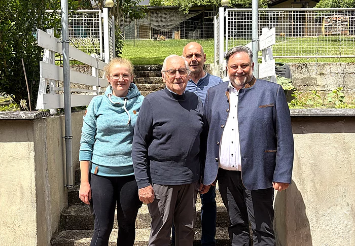 Vier Personen stehen bei sonnigem Wetter vor einer Treppe mit weißem Geländer und einem von Bäumen und Sträuchern umgebenen Grundstück, während im Vordergrund ein älterer Mann und ein Mann im dunklen Sakko sowie links eine junge Frau und dahinter ein weiterer Mann in die Kamera blicken.