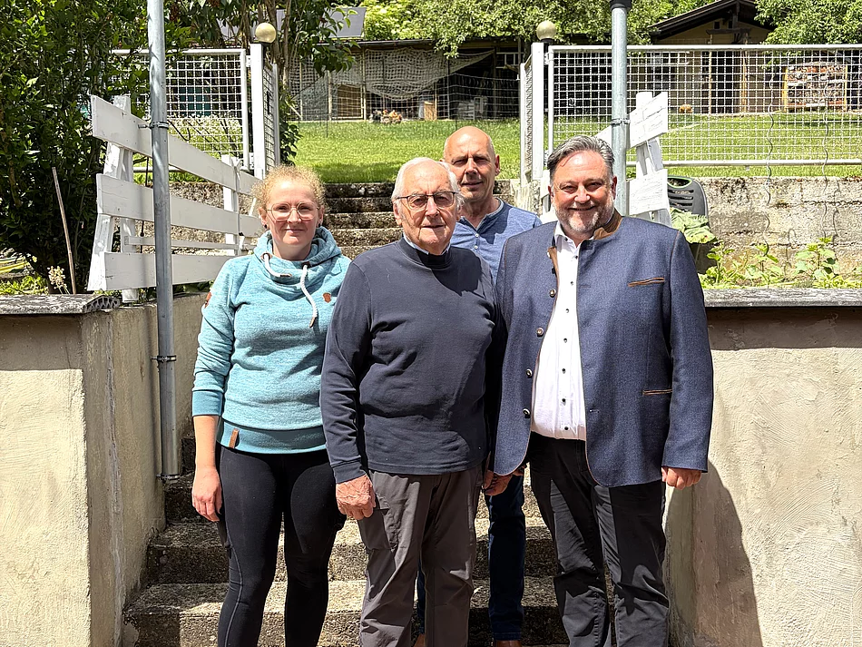 Vier Personen stehen bei sonnigem Wetter vor einer Treppe mit weißem Geländer und einem von Bäumen und Sträuchern umgebenen Grundstück, während im Vordergrund ein älterer Mann und ein Mann im dunklen Sakko sowie links eine junge Frau und dahinter ein weiterer Mann in die Kamera blicken.