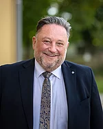 Ein Mann im Anzug mit Krawatte lächelt breit in die Kamera.