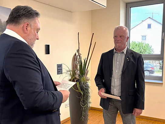 Ein Mann im Anzug liest aus einem Dokument vor, während ihm ein anderer Mann gegenübersteht und aufmerksam zuhört. Beide halten jeweils ein Blatt Papier. Die Situation wirkt wie ein offizieller Akt, etwa eine Vereidigung oder Ernennung. Im Raum sind eine Pflanze und ein Fenster mit Blick nach draußen zu sehen.
