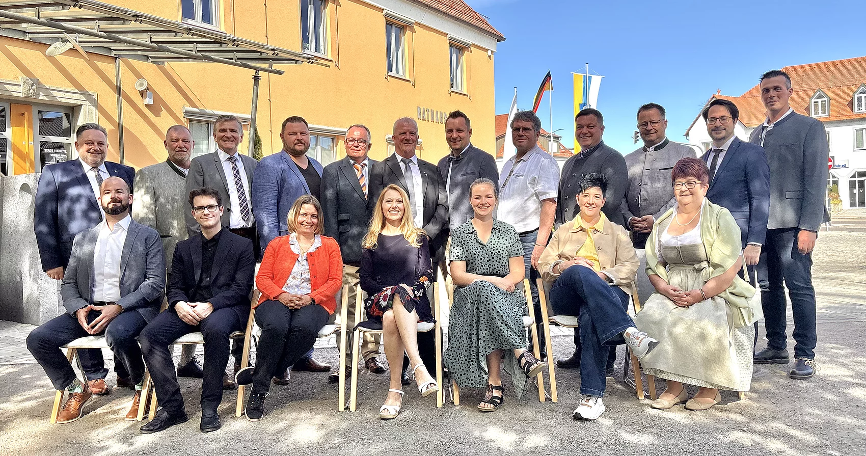 Eine größere Gruppe von Frauen und Männern sitzt und steht gemeinsam vor einem Gebäude und posiert bei sonnigem Wetter für ein Gruppenfoto.