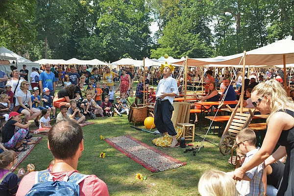 Ein Gaukler jongliert auf einem Teppich stehend umringt von Gästen und Kindern.