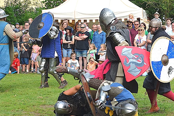 Mehrere Ritter in vollständigen Rüstungen kämpfen mit Schwertern und Schilden auf einer Wiese. Einer der Kämpfer liegt bereits am Boden, während die anderen den Kampf fortsetzen. Im Hintergrund verfolgen zahlreiche Besucherinnen und Besucher das Geschehen hinter einer Absperrung.