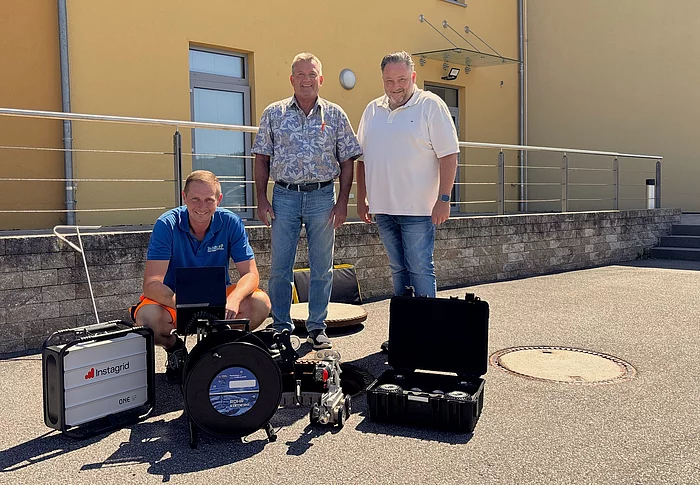 Ein Mitarbeiter des Klärwerks kniet vor einer neu angeschafften Kanal-Kamera mit Fahrwagensystem und Kontrolltechnik, während zwei Vertreter des Zweckverbands hinter ihm stehen. Die drei begutachten gemeinsam die moderne Ausrüstung, die künftig für die Inspektion von Abwasserkanälen eingesetzt wird.