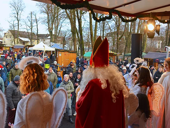 Das Bild zeigt eine festliche Szene auf einem Weihnachtsmarkt, bei der mehrere als Engel und Nikolaus verkleidete Personen von einer Bühne aus auf eine fröhliche Menschenmenge vor weihnachtlich geschmückten Buden blicken.