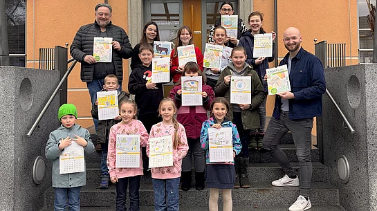 Eine Gruppe von Kindern und Erwachsenen steht auf der Treppe vor einem Gebäude und präsentiert selbst gestaltete Kalenderblätter, die sie hochhalten und stolz zeigen.
