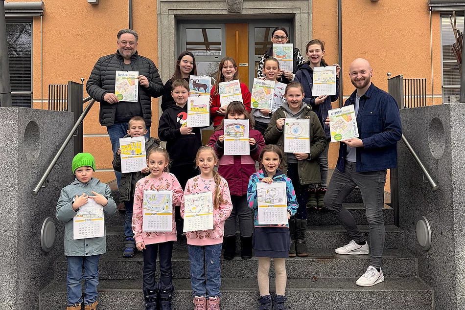 Eine Gruppe von Kindern und Erwachsenen steht auf der Treppe vor einem Gebäude und präsentiert selbst gestaltete Kalenderblätter, die sie hochhalten und stolz zeigen.