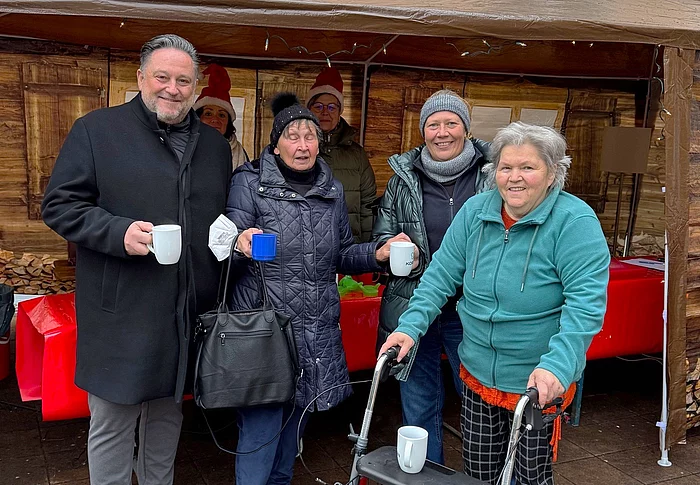 Mehrere Menschen stehen lächelnd mit warmen Getränken vor einer kleinen Weihnachtsmarktbude, während eine Frau mit Rollator im Vordergrund steht.