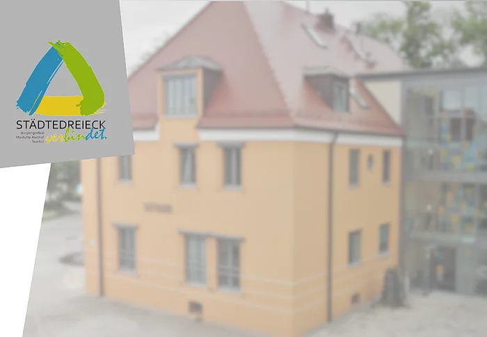 Grafik mit dem Logo des Städtedreiecks auf grauem Hintergrund. Im Hintergrund ist unscharf ein Gebäude mit gelber Fassade und modernem Glasanbau zu sehen. Die Grafik dient als Titelbild für eine Meldung über die Betriebsurlaube der Kleiderkammer im Städtedreieck und des Interkommunalen Recyclinghofs.