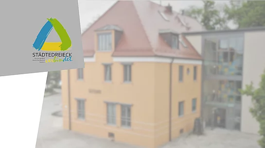 Grafik mit dem Logo des Städtedreiecks auf grauem Hintergrund. Im Hintergrund ist unscharf ein Gebäude mit gelber Fassade und modernem Glasanbau zu sehen. Die Grafik dient als Titelbild für eine Meldung über die Betriebsurlaube der Kleiderkammer im Städtedreieck und des Interkommunalen Recyclinghofs.