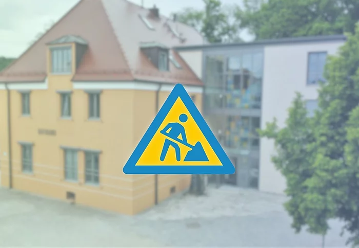 Im Vordergrund befindet sich ein großes, dunkelblaues Baustellensymbol – ein Dreieck mit einer stilisierten Person, die mit einer Schaufel arbeitet.  Der Hintergrund ist weichgezeichnet und zeigt das Rathaus von Teublitz in warmen Gelb- und Beigetönen.