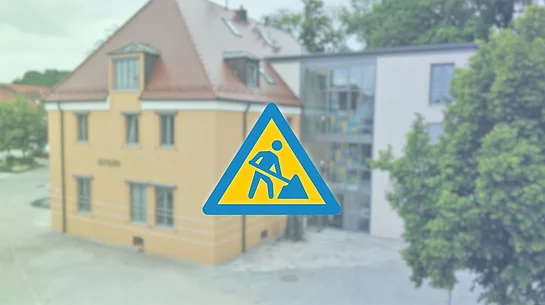 Im Vordergrund befindet sich ein großes, dunkelblaues Baustellensymbol – ein Dreieck mit einer stilisierten Person, die mit einer Schaufel arbeitet.  Der Hintergrund ist weichgezeichnet und zeigt das Rathaus von Teublitz in warmen Gelb- und Beigetönen.