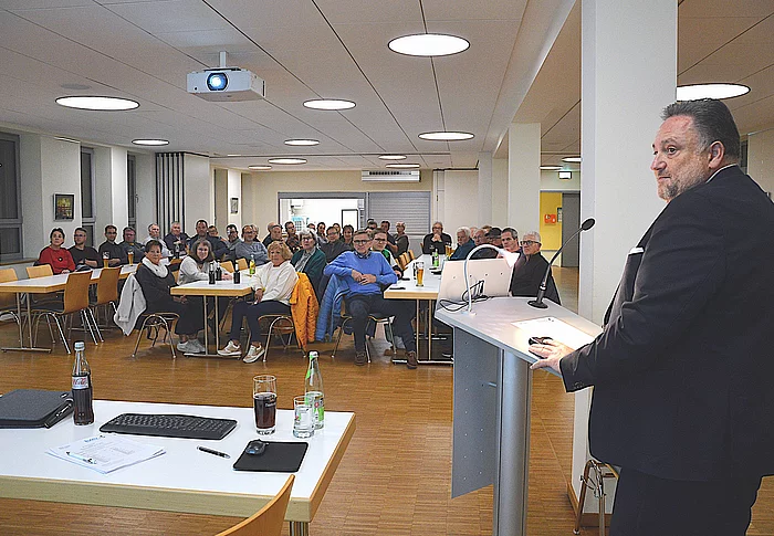 Das Bild zeigt eine Bürgerversammlung in einem hellen Saal, bei der ein Mann im Anzug an einem Rednerpult spricht, während zahlreiche Zuhörer aufmerksam an Tischen sitzen und ihm zuhören.