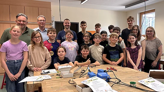 Eine Gruppe von Kindern und Jugendlichen steht gemeinsam mit mehreren Erwachsenen in einem Werkraum und lächelt in die Kamera. Auf den Arbeitstischen liegen Werkzeuge, Kabel und Materialien, was auf eine praktische Technik- oder Bastelaktivität hindeutet.