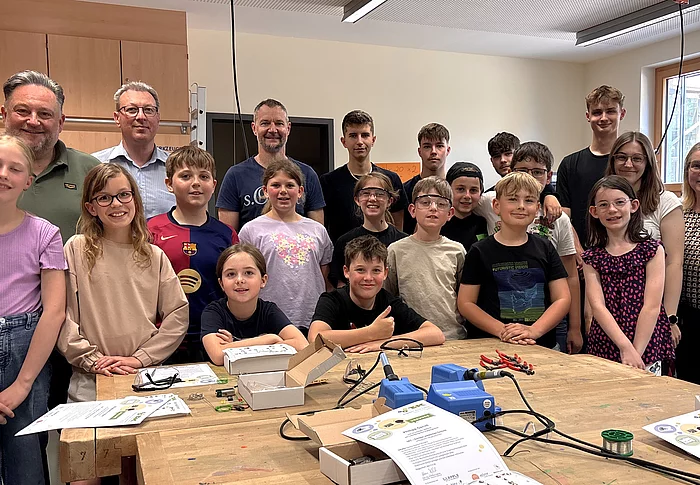 Eine Gruppe von Kindern und Jugendlichen steht gemeinsam mit mehreren Erwachsenen in einem Werkraum und lächelt in die Kamera. Auf den Arbeitstischen liegen Werkzeuge, Kabel und Materialien, was auf eine praktische Technik- oder Bastelaktivität hindeutet.