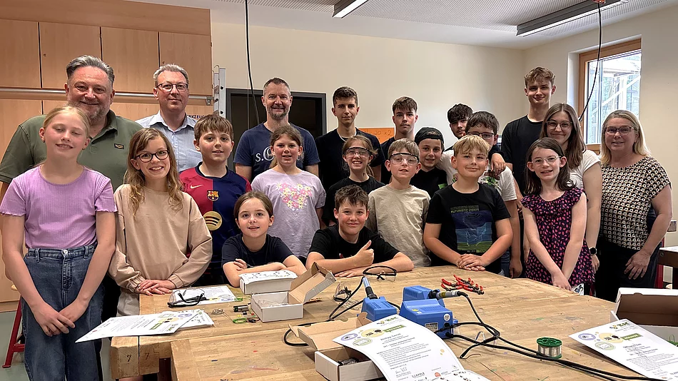 Eine Gruppe von Kindern und Jugendlichen steht gemeinsam mit mehreren Erwachsenen in einem Werkraum und lächelt in die Kamera. Auf den Arbeitstischen liegen Werkzeuge, Kabel und Materialien, was auf eine praktische Technik- oder Bastelaktivität hindeutet.