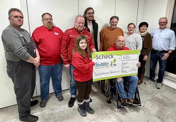 Auf dem Foto steht eine Gruppe von Menschen zusammen, während ein Mädchen und ein Mann im Rollstuhl symbolisch einen großen Spendenscheck über 600 Euro präsentieren.