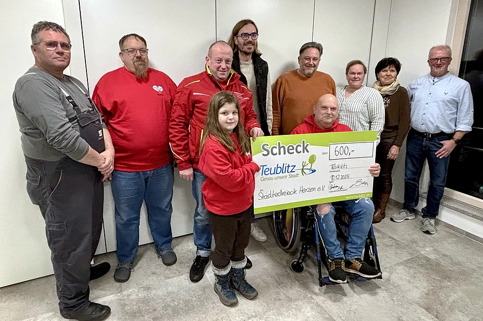 Auf dem Foto steht eine Gruppe von Menschen zusammen, während ein Mädchen und ein Mann im Rollstuhl symbolisch einen großen Spendenscheck über 600 Euro präsentieren.
