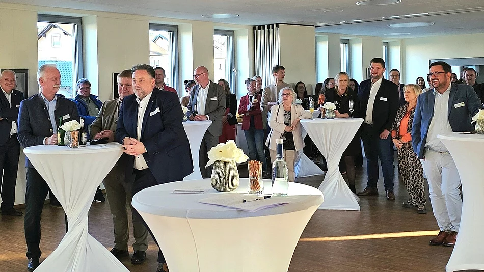 Eine Gruppe von Menschen steht bei einem Empfang in einem hellen Raum an Stehtischen zusammen und hört aufmerksam zu.