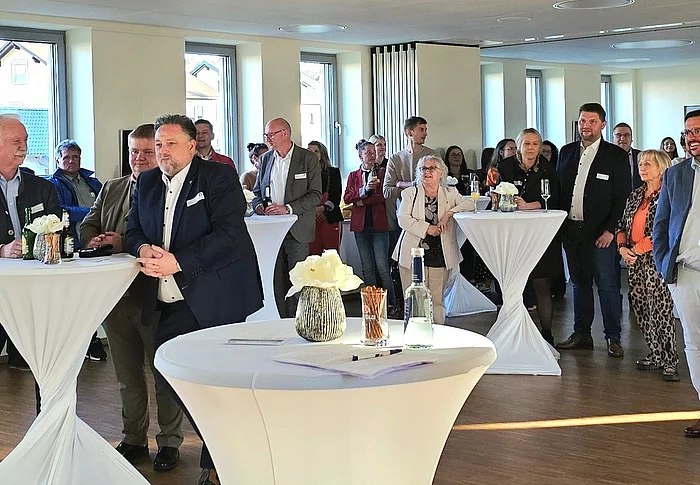 Eine Gruppe von Menschen steht bei einem Empfang in einem hellen Raum an Stehtischen zusammen und hört aufmerksam zu.