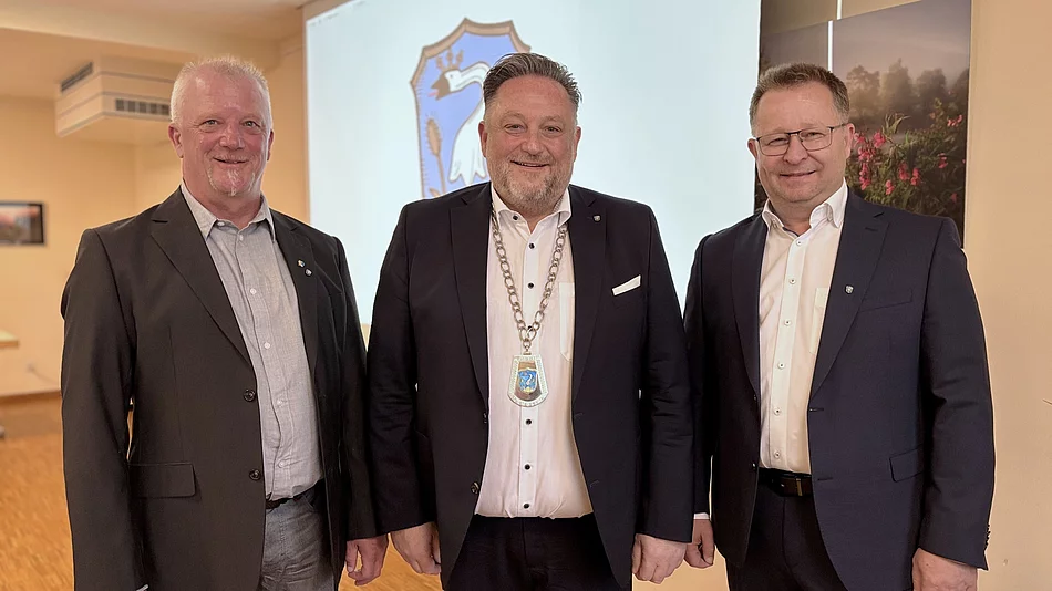 Drei Männer stehen nebeneinander und blicken in die Kamera. Der Mann in der Mitte trägt eine Amtskette und steht damit erkennbar im Mittelpunkt. Alle drei sind formell gekleidet und lächeln. Im Hintergrund ist ein Wappen zu sehen, das auf den offiziellen Rahmen der Aufnahme hinweist.
