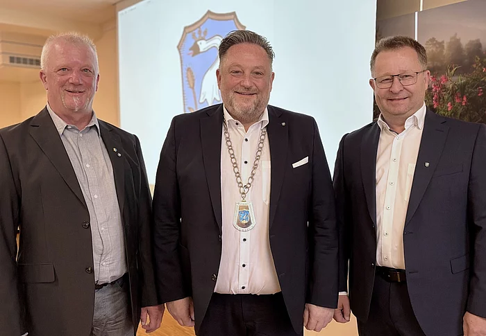 Drei Männer stehen nebeneinander und blicken in die Kamera. Der Mann in der Mitte trägt eine Amtskette und steht damit erkennbar im Mittelpunkt. Alle drei sind formell gekleidet und lächeln. Im Hintergrund ist ein Wappen zu sehen, das auf den offiziellen Rahmen der Aufnahme hinweist.