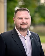 Ein Mann mit kurzem Haar und Bart trägt ein Sakko und schaut neutral.