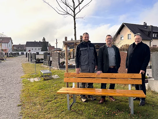 Drei Männer stehen lächelnd hinter einer neuen Holzbank auf einem Friedhof, umgeben von Grabsteinen, Rasenflächen und Häusern im Hintergrund unter leicht bewölktem Himmel.