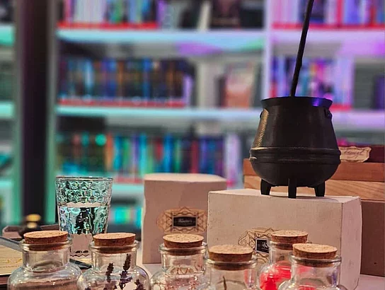 Auf einem Tisch stehen mehrere kleine Glasfläschchen mit getrockneten Pflanzen und Zutaten sowie ein kleiner schwarzer Kessel mit Rührstab. Im Hintergrund sind unscharf beleuchtete Bücherregale zu erkennen. Die Dekoration vermittelt eine magische und geheimnisvolle Stimmung.