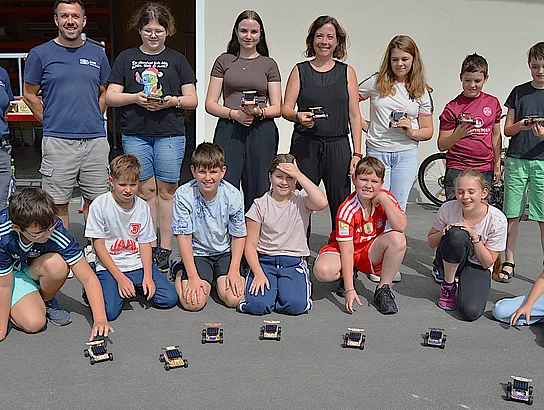 Eine Gruppe aus Kindern, Jugendlichen und Erwachsenen steht im Freien vor einem Gebäude. Mehrere Teilnehmende halten selbst gebaute kleine Fahrzeuge in den Händen, weitere Fahrzeuge stehen vor der Gruppe auf dem Boden.