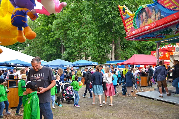Viele Menschen gehen zwischen bunt beleuchteten Buden und Fahrgeschäften über einen Volksfestplatz unter großen Bäumen.