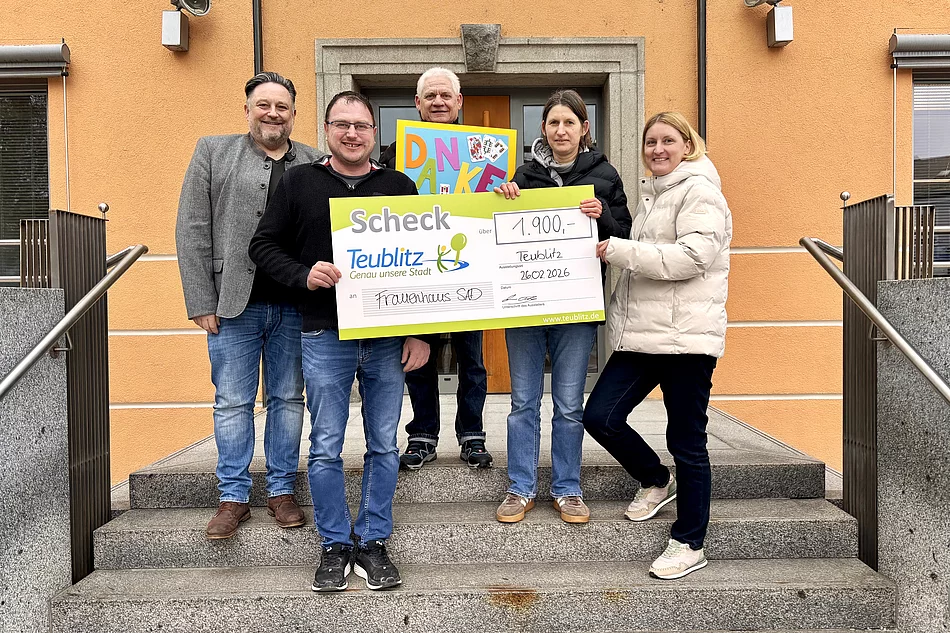 Mehrere Personen stehen vor einem Gebäude auf einer Treppe und halten gemeinsam einen großen symbolischen Spendenscheck sowie ein buntes „Danke“-Plakat in die Höhe.