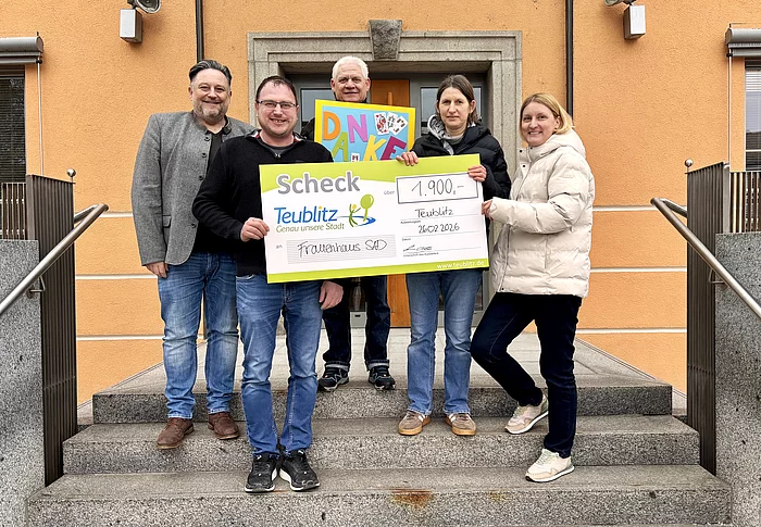 Mehrere Personen stehen vor einem Gebäude auf einer Treppe und halten gemeinsam einen großen symbolischen Spendenscheck sowie ein buntes „Danke“-Plakat in die Höhe.