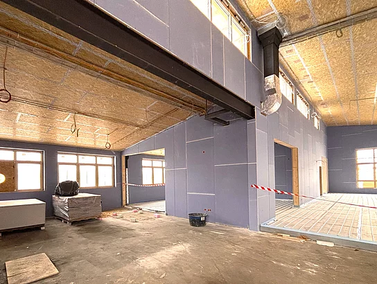 Ein leerer, noch unfertiger Innenraum mit hohen Decken, sichtbaren Leitungen und Baumaterialien zeigt den Baufortschritt eines Gebäudes.